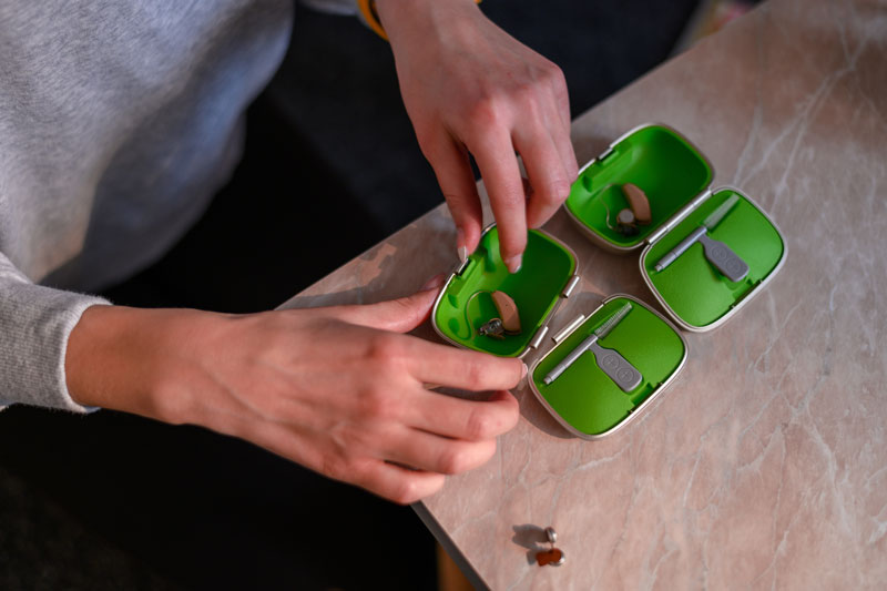 A case containing two hearing aids is visible with a pair of hands about to select one.