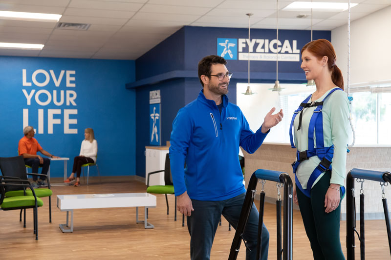 A therapist is extending their hand to assist a red-headed woman in maintaining her balance on the equipment.